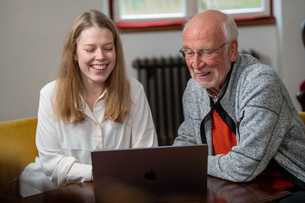 Eine junge Frau und ein älterer Herr sitzen lachend vor einem Laptop.