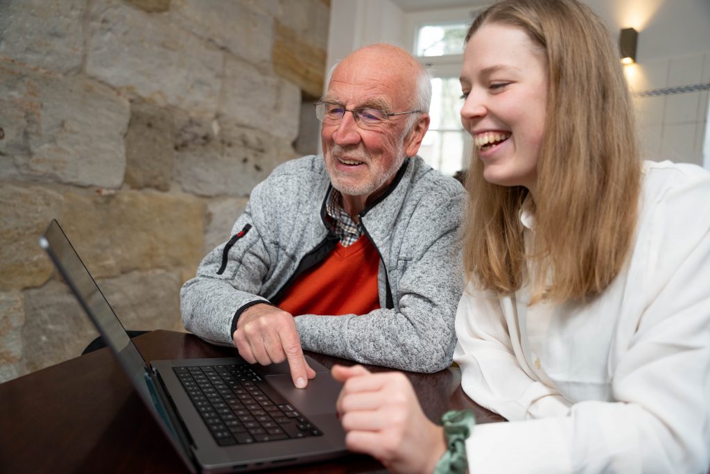 Eine junge Frau und ein älterer Herr sitzen lachend vor einem Laptop.