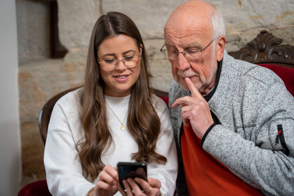Eine junge Frau mit runder Brille und weißem Pullover zeigt einem älteren Herren in einem grauen Pullover Dinge am Handy. Die junge Frau lacht, der ältere Herr schaut etwas überlegend und hält sich den Finger an den Mund.